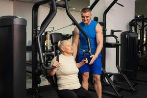 een knap jong trainer Mens helpt een oud vrouw trein sport- in Sportschool. gelukkig mensen glimlach, genieten leven. actief, gezond levensstijl, senior. foto