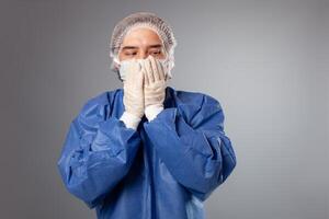 een knap mannetje chirurg is een dokter Aan een grijs achtergrond in de studio. een Mens in een gewaad, een medisch masker, een hoed. leeg ruimte. een Mens covers zijn mond met zijn handen, doet niet willen naar zeggen iets. foto