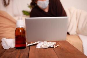 een ziek Mens in een medisch masker tegen de achtergrond van tabletten. huis quarantaine, coronavirus, een Mens leugens Aan een bed met een laptop. foto