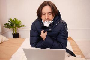 een ziek Mens bevroor Bij huis. zijn heel koud. een Mens zit in buitenste kleding achter een laptop vervelend een masker. huis quarantaine in winter. coronavirus. een Mens met pillen in zijn tanden. foto