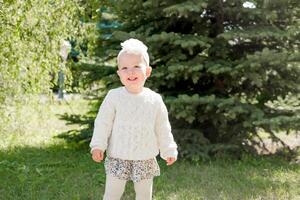 schattig blond meisje in een jurk in pastel kleuren en wit panty tegen de backdrop van de park. 1 jaar oud baby leert naar wandelen foto