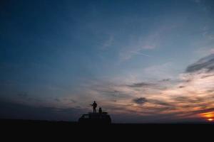 silhouet van een man met een gitaar en een vrouw in een hoedenstandaard op het dak van een auto op de achtergrond van de zonsondergang foto