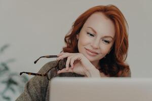 roodharig professioneel vrouw Holding bril, op zoek Bij laptop scherm met een contemplatief glimlach foto