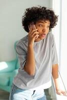 Afrikaanse Amerikaans meisje pratend Aan smartphone Bij huis binnen. jong vrouw met cel telefoon chatten met vrienden. glimlachen tiener meisje maken beantwoorden telefoontje door mobiele telefoon. dame hebben gesprek door mobiel foto