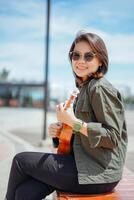 spelen ukulele van jong mooi Aziatisch vrouw vervelend jasje en zwart jeans poseren buitenshuis foto