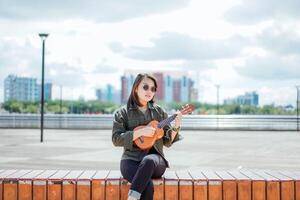 spelen ukulele van jong mooi Aziatisch vrouw vervelend jasje en zwart jeans poseren buitenshuis foto