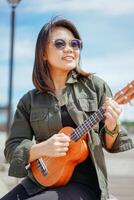 spelen ukulele van jong mooi Aziatisch vrouw vervelend jasje en zwart jeans poseren buitenshuis foto