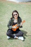 spelen ukulele van jong mooi Aziatisch vrouw vervelend jasje en zwart jeans poseren buitenshuis foto