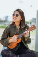 spelen ukulele van jong mooi Aziatisch vrouw vervelend jasje en zwart jeans poseren buitenshuis foto