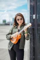 spelen ukulele van jong mooi Aziatisch vrouw vervelend jasje en zwart jeans poseren buitenshuis foto