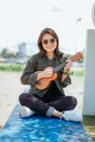 spelen ukulele van jong mooi Aziatisch vrouw vervelend jasje en zwart jeans poseren buitenshuis foto