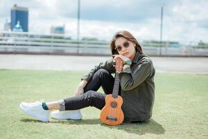 spelen ukulele van jong mooi Aziatisch vrouw vervelend jasje en zwart jeans poseren buitenshuis foto