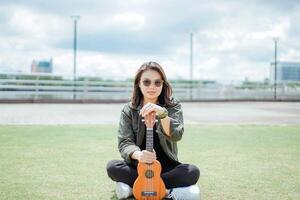 spelen ukulele van jong mooi Aziatisch vrouw vervelend jasje en zwart jeans poseren buitenshuis foto
