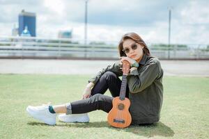 spelen ukulele van jong mooi Aziatisch vrouw vervelend jasje en zwart jeans poseren buitenshuis foto