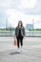 spelen ukulele van jong mooi Aziatisch vrouw vervelend jasje en zwart jeans poseren buitenshuis foto