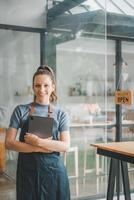 glimlachen vrouw werknemer in een denim schort gebruik makend van een digitaal tablet in een knus cafe met een 'open' teken in de achtergrond. foto