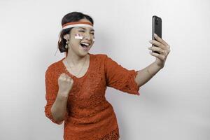 een jong Aziatisch vrouw met een gelukkig geslaagd uitdrukking terwijl Holding haar telefoon en vervelend rood kebaya, vlag hoofdband geïsoleerd door wit achtergrond. Indonesië's onafhankelijkheid dag concept. foto