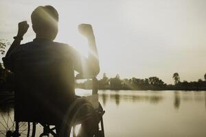 silhouet jong gehandicapt Mens zien rivier- achtergrond.he is verhogen een hand- naar en zittend Aan rolstoel.wanhoop,eenzaam,hoop.foto concept depressie en geduldig. foto