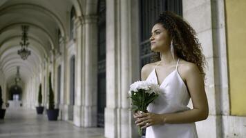 mooi zwart vrouw in elegant wit jurk. actie. aantrekkelijk vrouw in wit jurk met boeket Bij oud gebouw. aantrekkelijk jong vrouw is aan het wachten voor vriendje met bloemen foto