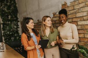 drie jong bedrijf Dames met digitaal tablet staand door de steen muur in de industrieel stijl kantoor foto