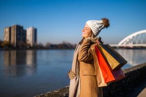 gelukkig vrouw in warm kleding met boodschappen doen Tassen geniet staand door rivier- Aan zonnig winter dag. foto