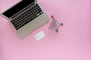 computer laptop met blanco credit kaart en mini boodschappen doen kar of trolley Aan roze achtergrond foto