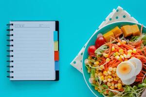 vers salade groente met gekookt kip ei en blanco spiraal notitieboekje Aan blauw achtergrond foto