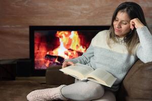 vrouw geniet lezing een boek van haar sofa Bij huis. foto