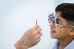 detailopname van optometrist aan het doen zicht testen voor kind geduldig in kliniek, Indisch kind kiezen bril in optiek op te slaan, jongen aan het doen oog test controle examen met optometrist in optisch winkel foto