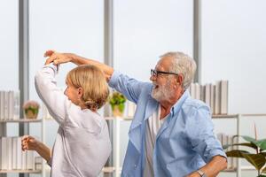 senior paar vrouw en man dansen naar muziek- samen in leven kamer, gepensioneerd Mens en vrouw hebben pret vrij tijd samen Bij huis foto