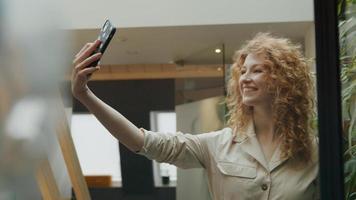 jonge blanke vrouw houdt mobiele telefoon in de lucht, past haar haar aan, lacht naar de telefoon, beweegt de telefoon voor haar, kijkt naar het scherm foto