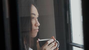 jonge vrouw van gemengd ras loopt met beker met beide handen vast, loopt naar het raam, neemt een slokje, kijkt contemplatief door raam foto