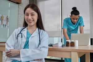 portret van mooie vrouwelijke arts van Aziatische etniciteit in uniform met stethoscoop. glimlach en kijk naar de camera in een ziekenhuiskliniek, mannelijke partner die achter haar werkt, twee professionele personen.