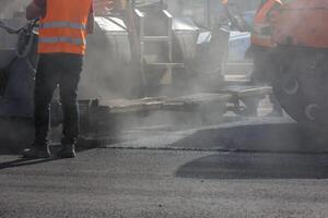 mannen werken met asfalteren bestratingsafwerkmachine machine gedurende weg straat repareren werken Bij dag licht met rook en stoom- in de lucht foto