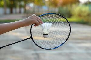 hand- Holding een wit shuttle in voorkant van een badminton racket, groen achtergrond van bomen. zacht en selectief focus foto