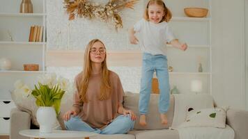 bedachtzaam kalmte Kaukasisch moeder in bril mediteren zittend Aan sofa terwijl actief luidruchtig kind dochter jumping Aan bank. vredig mam ontspannende in yoga meditatie Bij huis ondeugend weinig kind meisje springen foto