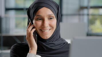 detailopname jong glimlachen Arabisch vrouw secretaris in hijab pratend Aan telefoon in kantoor vrouw verkoop middel raadplegen cliënt afgelegen antwoorden telefoontje gelukkig zakenvrouw genieten van mobiel gesprek gebruik makend van smartphone foto
