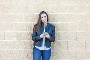 glimlachen vrouw op zoek verder Holding een smartphone staand leunend Aan een muur foto