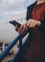 jong Aziatisch vrouw in Internationale luchthaven, gebruik makend van mobiel smartphone en controle vlucht Bij de vlucht informatie bord foto