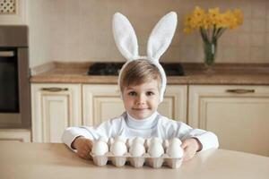 schattig Kaukasisch kind vervelend konijn oren en Holding reeks van wit biologisch chiken eieren, voorbereidingen treffen naar Pasen. weinig jongen zittend Bij tafel en op zoek Bij camera. foto