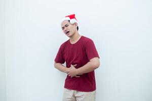 jong Aziatisch Mens vervelend een de kerstman claus hoed uitdrukken maagpijn door Holding zijn maag in pijn geïsoleerd door een wit achtergrond voor zichtbaar communicatie foto