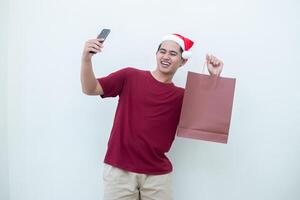 jong Aziatisch Mens vervelend een de kerstman claus hoed Holding een smartphone en een boodschappen doen zak met uitdrukkingen van glimlach, schok, en verrassing, geïsoleerd tegen een wit achtergrond voor zichtbaar communicatie foto