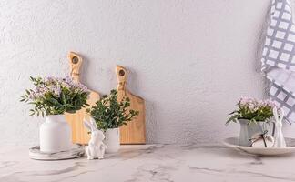 voorjaar bloemen in vaas en kruik Aan keuken aanrecht met Pasen decoraties. snijdend planken, handdoeken. Pasen konijn beeldjes. foto