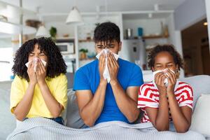 portret van ziek jong zwart familie van drie mensen blazen vloeibaar neuzen terwijl zittend samen Aan de sofa met servetten en gedekt met deken. foto