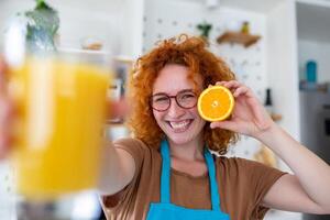 foto van schattig Kaukasisch vrouw glimlachen en Holding twee oranje onderdelen terwijl Koken groente salade in keuken interieur Bij huis