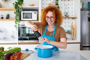 schattig jong vrouw Koken en toevoegen kruid naar maaltijd, lachend en uitgeven tijd in de keuken foto