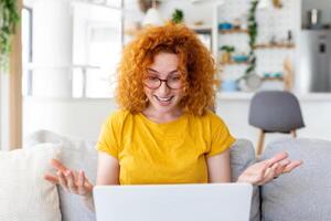 gelukkig jong vrouw hebben pret aan het doen video telefoontje gebruik makend van laptop in haar huis, golvend hand- video conferentie roeping Aan laptop computer zitten Aan sofa afstand leren zoom online virtueel vergadering Bij huis. foto