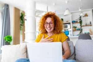 gelukkig jong vrouw hebben pret aan het doen video telefoontje gebruik makend van laptop in haar huis, golvend hand- video conferentie roeping Aan laptop computer zitten Aan sofa afstand leren zoom online virtueel vergadering Bij huis. foto