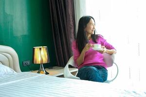 portret van mooi jong Aziatisch vrouw Holding kop van ochtend- koffie in wit slaapkamer, Vrolijk, vrolijk, ontspannende in zomer. Thais Aziatisch bedenken ideeën, wit slaapkamer foto