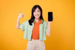 vrolijk Aziatisch vrouw Jaren 30 vervelend oranje en groen shirt. points vinger naar smartphone, symboliseert connectiviteit en digitaal machtiging. technologie en modern levensstijl Aan een levendig geel achtergrond. foto